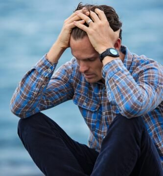 A man in a plaid shirt sits by the water looking distressed, symbolizing stress.