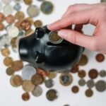 Close-up of a hand inserting a coin into a black piggy bank with scattered coins on a white background.