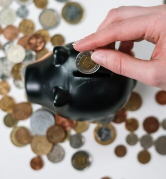 Close-up of a hand inserting a coin into a black piggy bank with scattered coins on a white background.