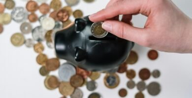 Close-up of a hand inserting a coin into a black piggy bank with scattered coins on a white background.